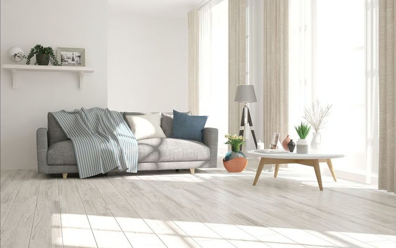 Staged living room with a gray sofa, blue cushions, pale wood floors, and tall sunlit windows.