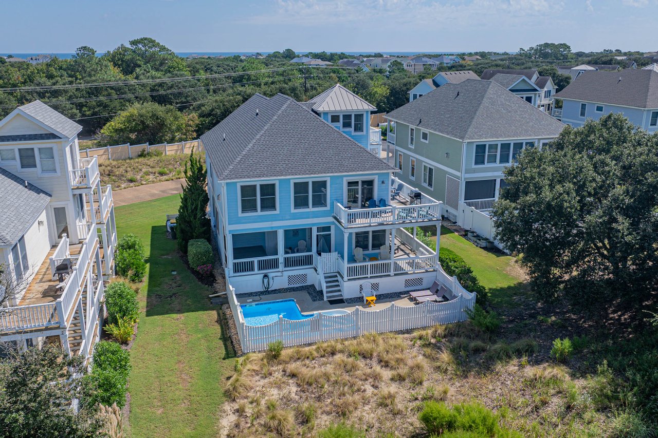 Blue coastal house with a fenced pool, neighboring homes and ocean in the distance.