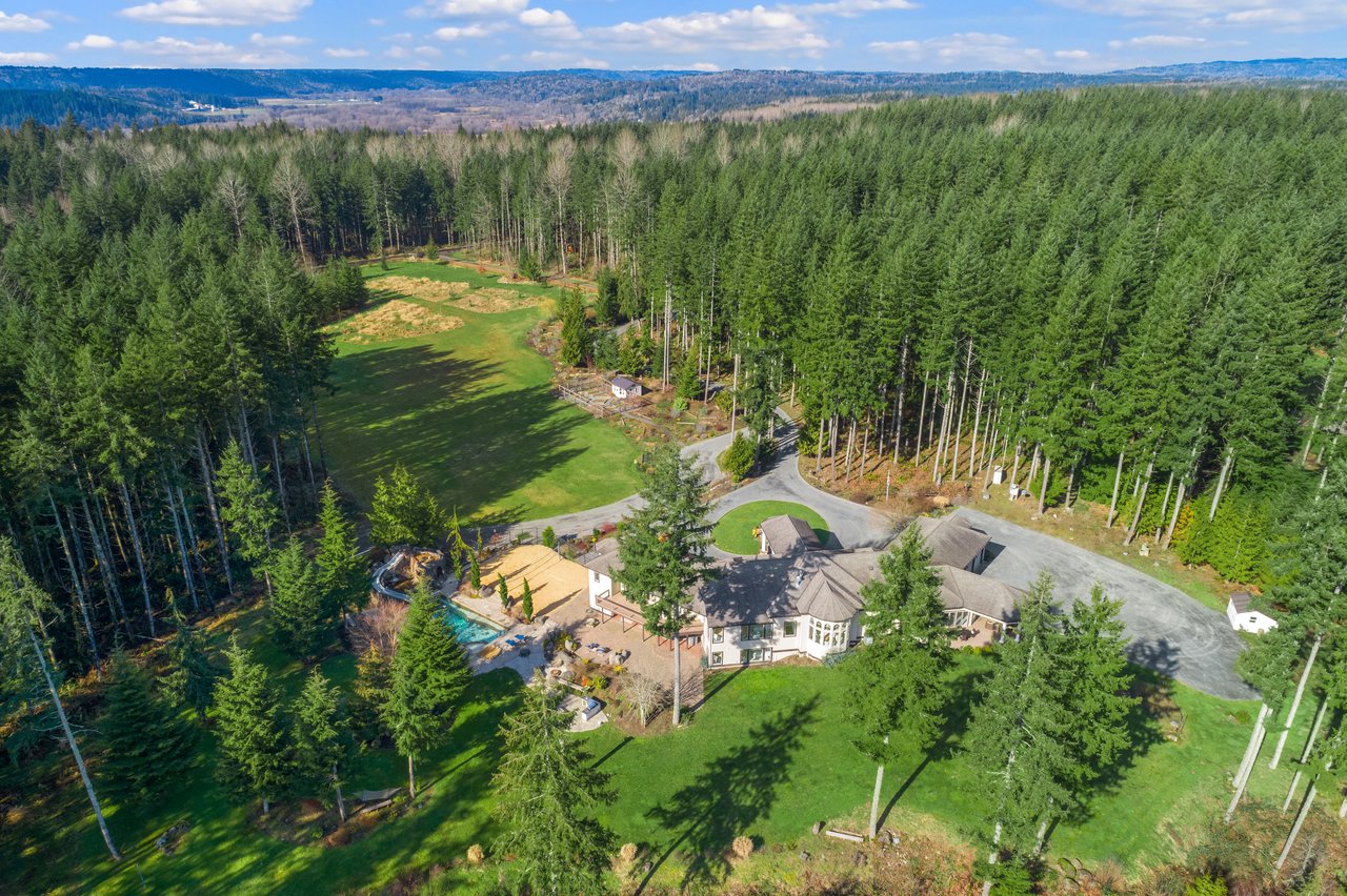 Aerial view of a home amid dense forest, with broad lawns, a swimming pool, and a curved driveway.