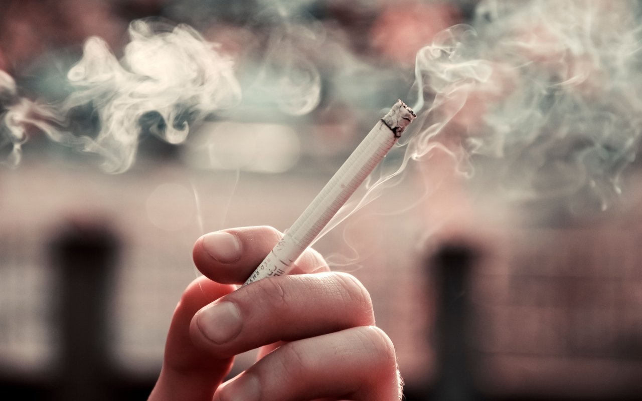 A hand holds a lit cigarette as smoke curls through the air.