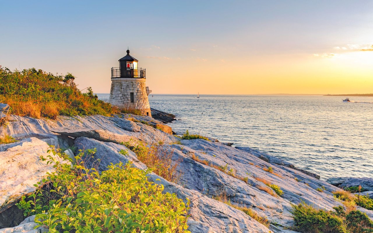 Lighthouse on Newport’s rocky coast at sunset, with boats visible offshore.