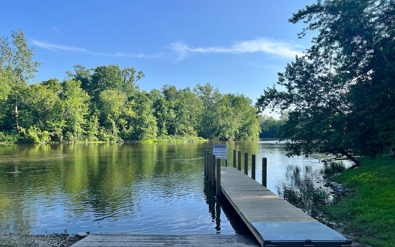 Wooden dock extending into a tree-lined river, with a shallow-water warning sign.