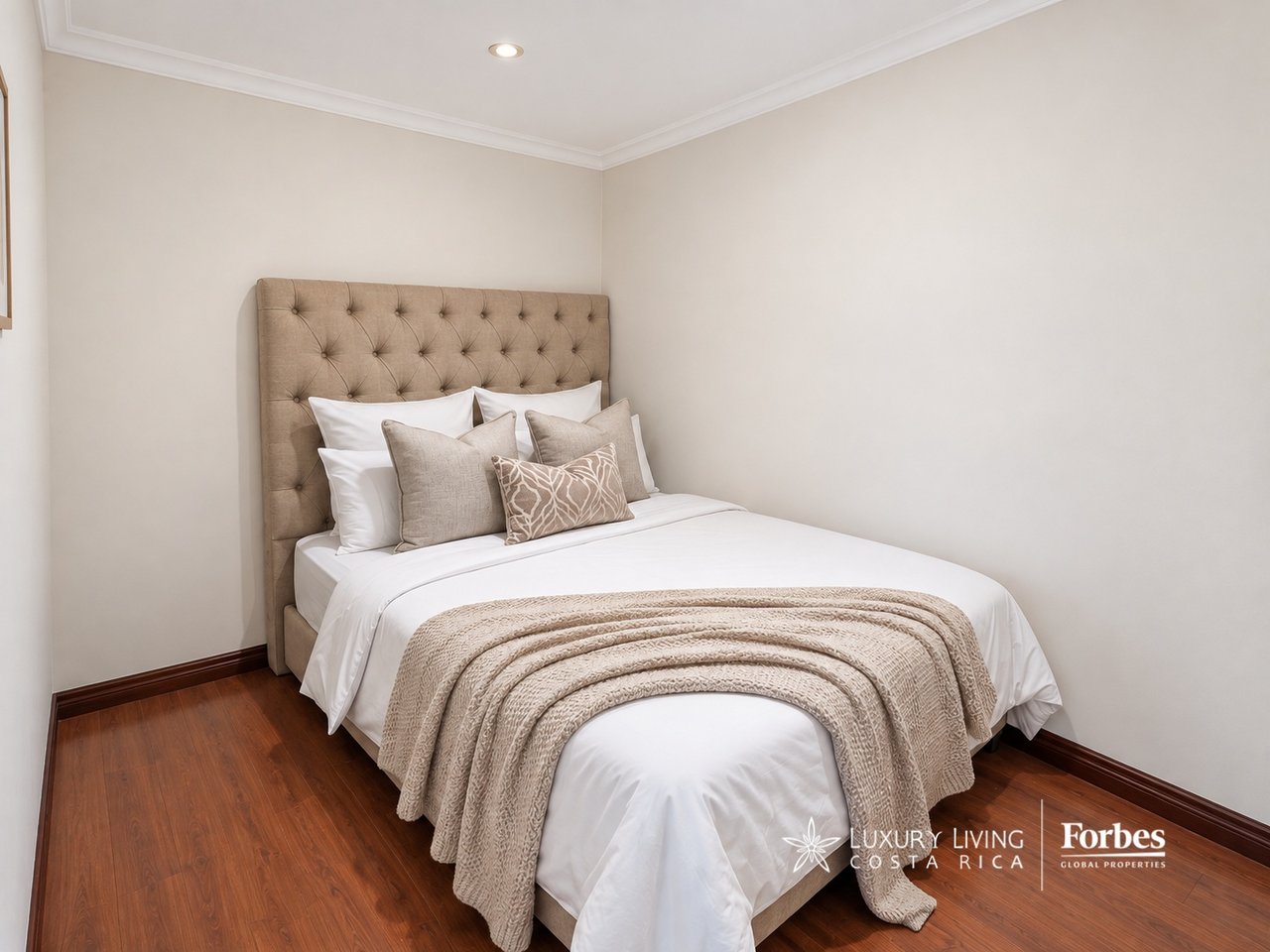 Bedroom with a white bed, tufted beige headboard, neutral pillows, and reddish-brown wood floors.