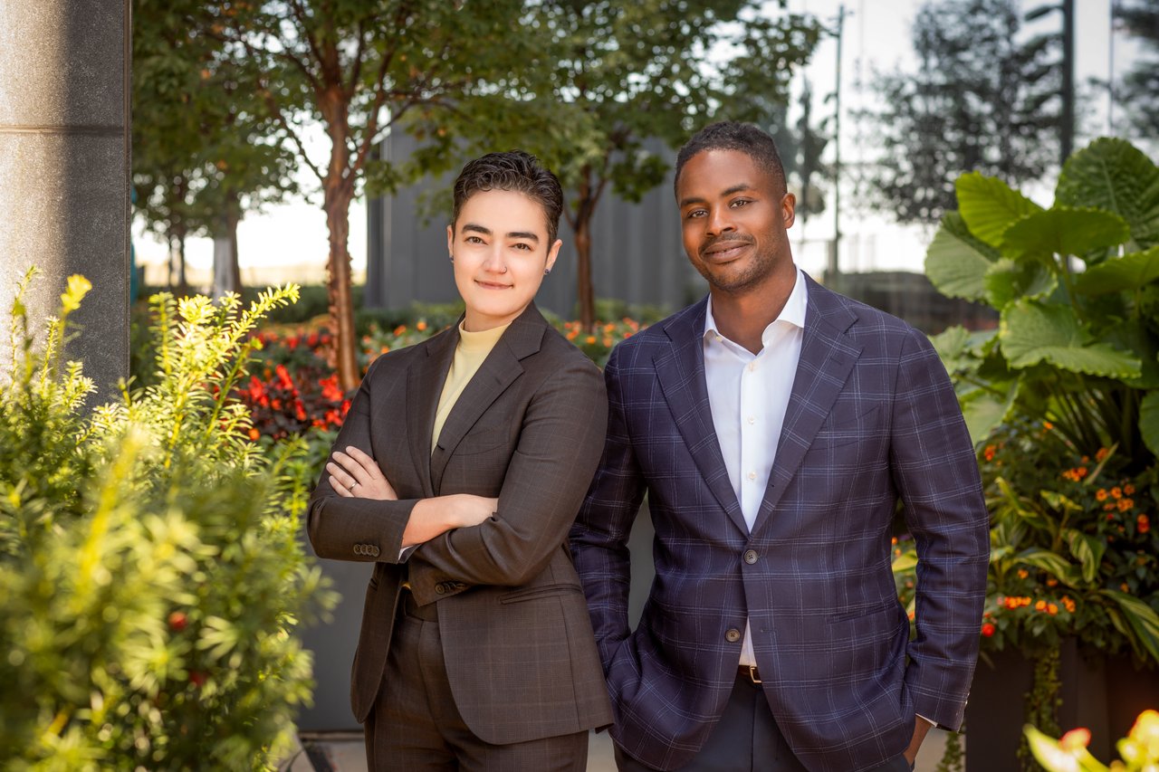 Two real estate agents in business attire pose among landscaped greenery.