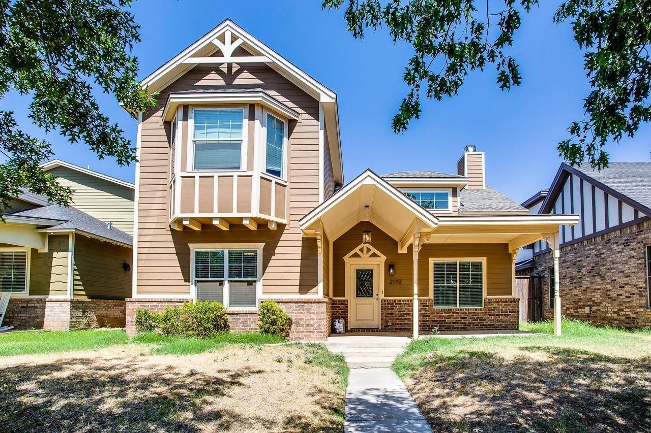 Two-story tan house with white-trimmed gables, brick base, covered entrance, and leafy trees framing the lawn.