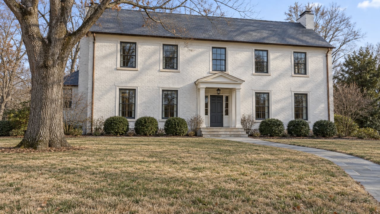 A painted-brick house with a slate roof, broad winter lawn, bare white oak, and stone entry walk in midday sunlight.