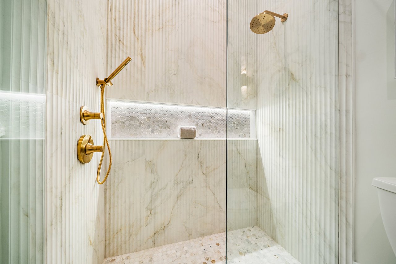 Walk-in shower with white marble tile, gold fixtures, a recessed niche, and a pebble mosaic floor.