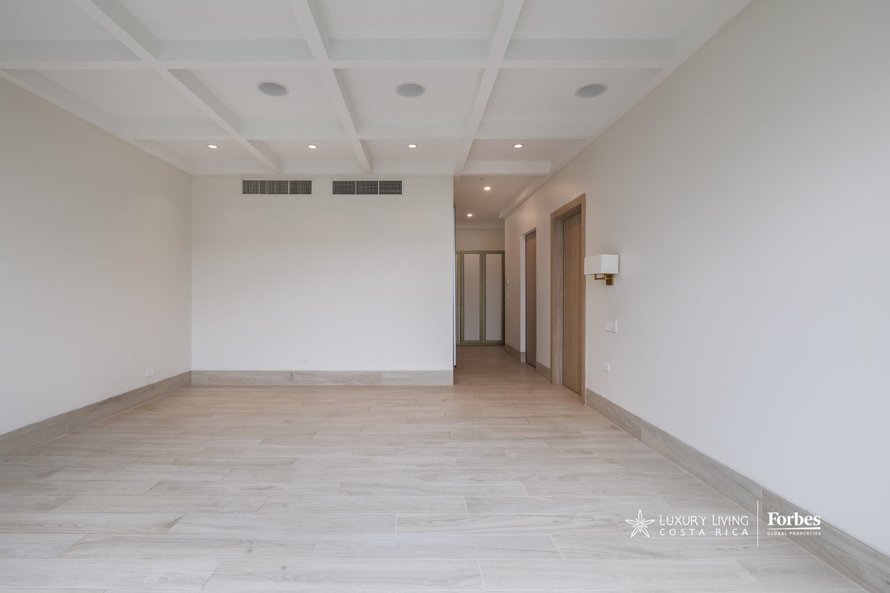 Empty apartment room with pale tile flooring, a coffered ceiling, and a hallway with wooden doors.