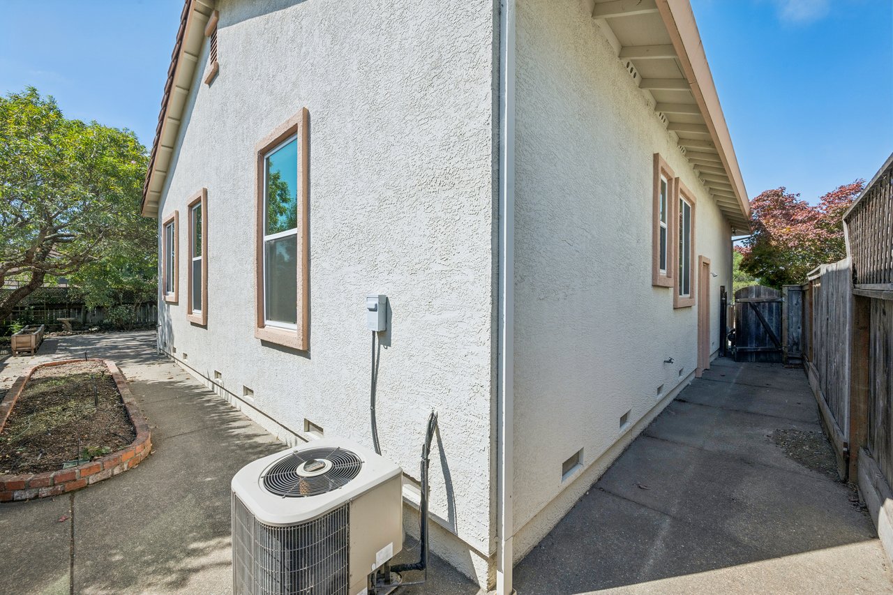 Stucco house side exterior with tan-trimmed windows, a concrete walkway, and an outdoor air-conditioning unit.