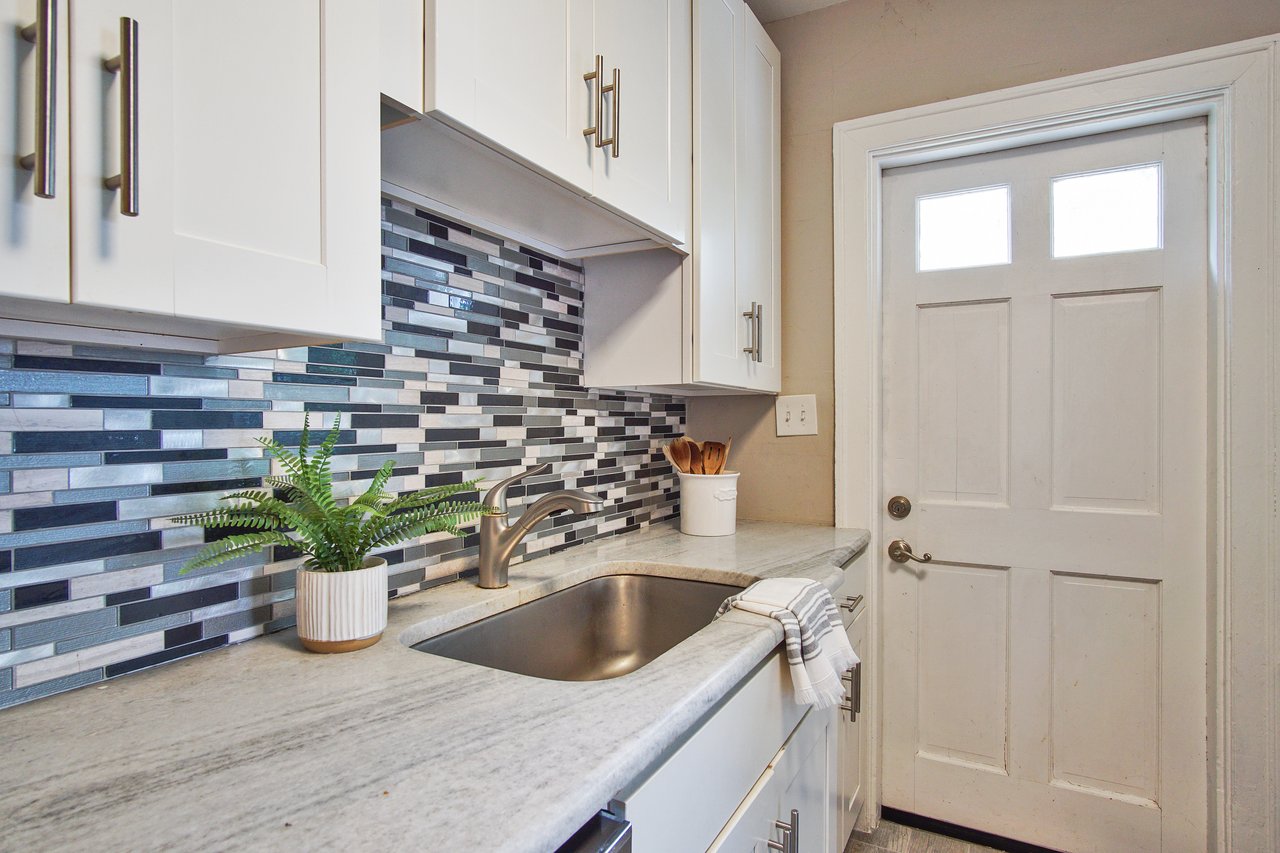 Modern kitchen with white cabinets and a sleek gray marble countertop. A stainless-steel sink, potted plant, and white door complete the clean, fresh look.