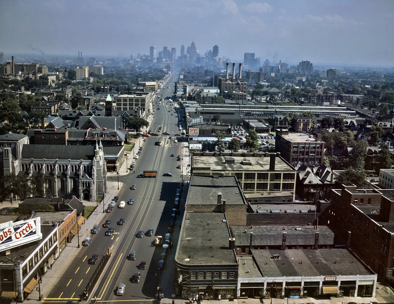 Historic Detroit avenue lined with commercial buildings, with traffic and the downtown skyline in the distance.