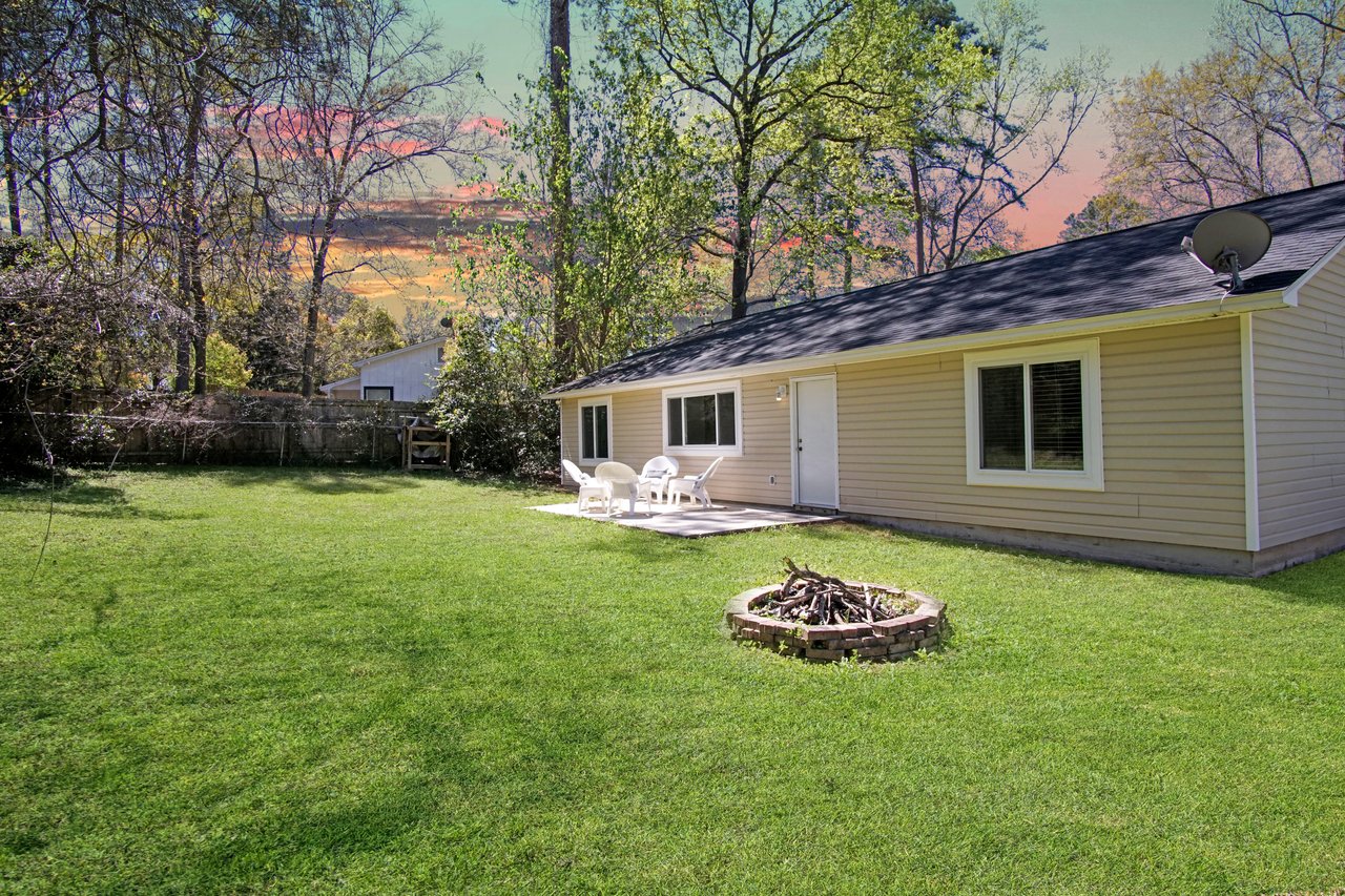 Small beige house with a dark roof, surrounded by green grass and trees. A patio with white chairs and a fire pit add a cozy, peaceful vibe at sunset.