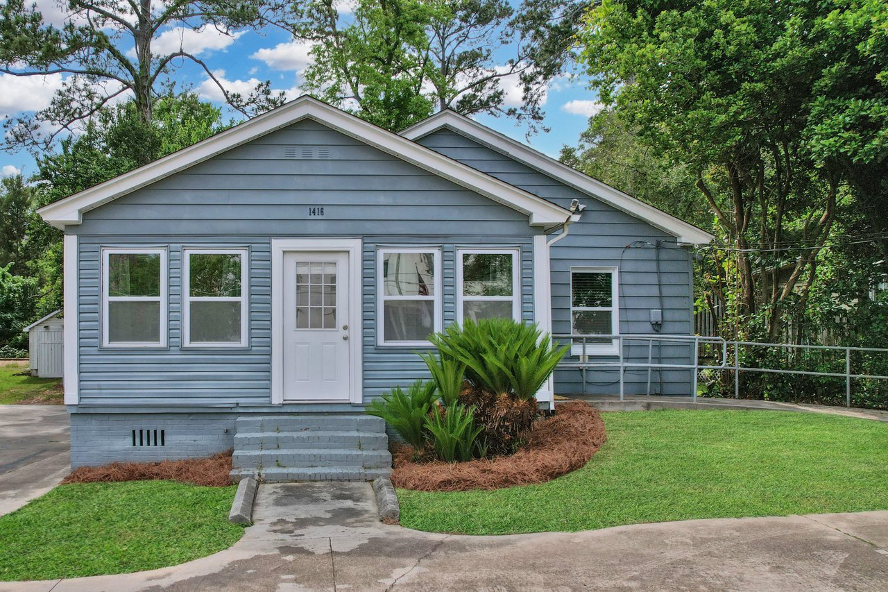 A small, single-story blue house with white trim and a front door, surrounded by lush greenery. Steps lead to the entrance, with neat landscaping in front.