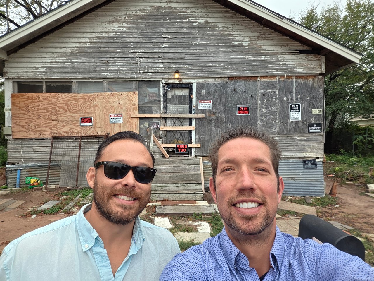 Two men pose in front of a boarded, weathered wooden house.