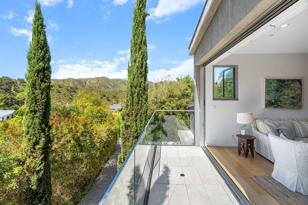 Balcony with glass railing overlooking cypress trees and hills, beside a bright living room.