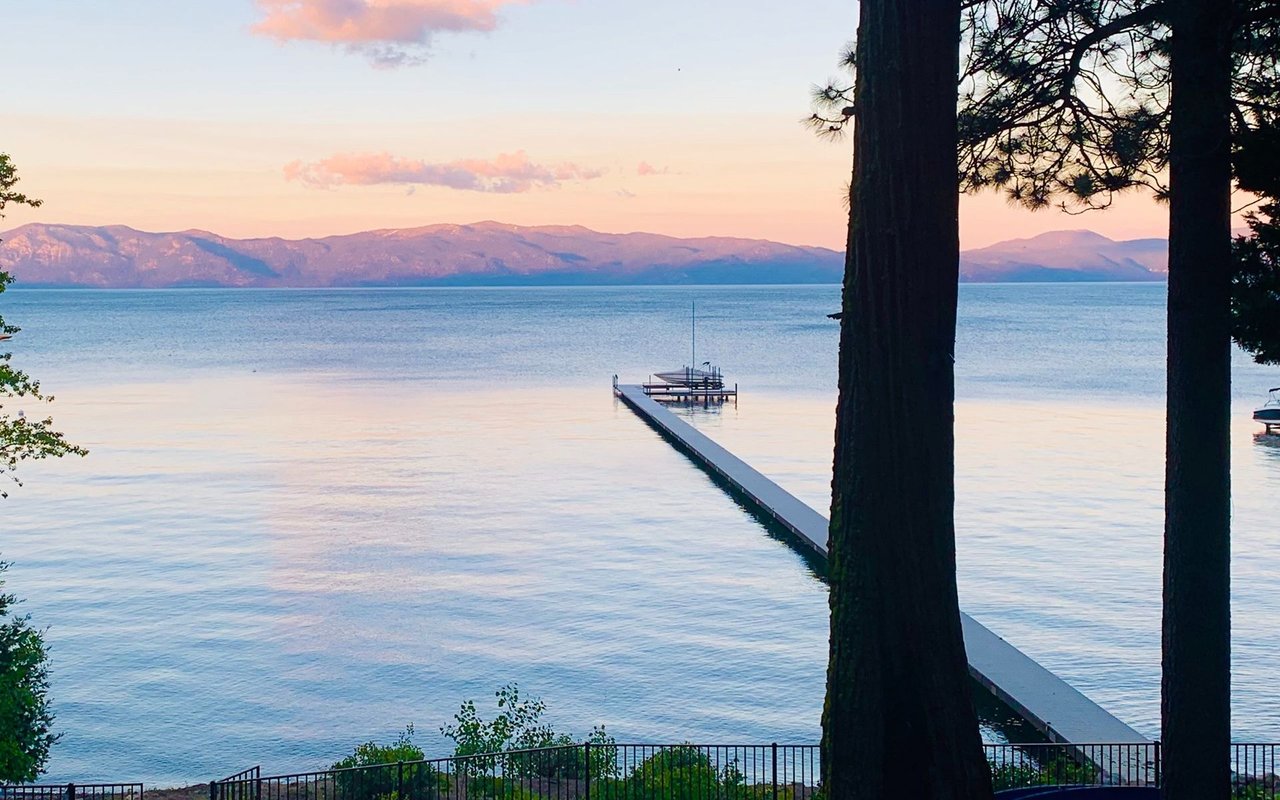 Lake Tahoe at sunset, framed by pine trees, with a long dock reaching toward distant mountains.