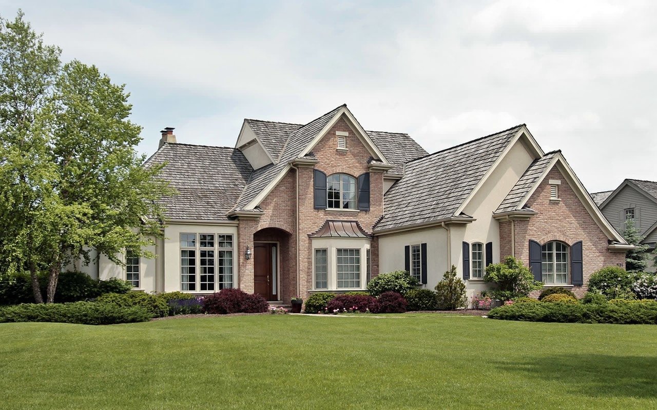 A large, multi-gabled brick and stucco luxury home fronted by a manicured green lawn.