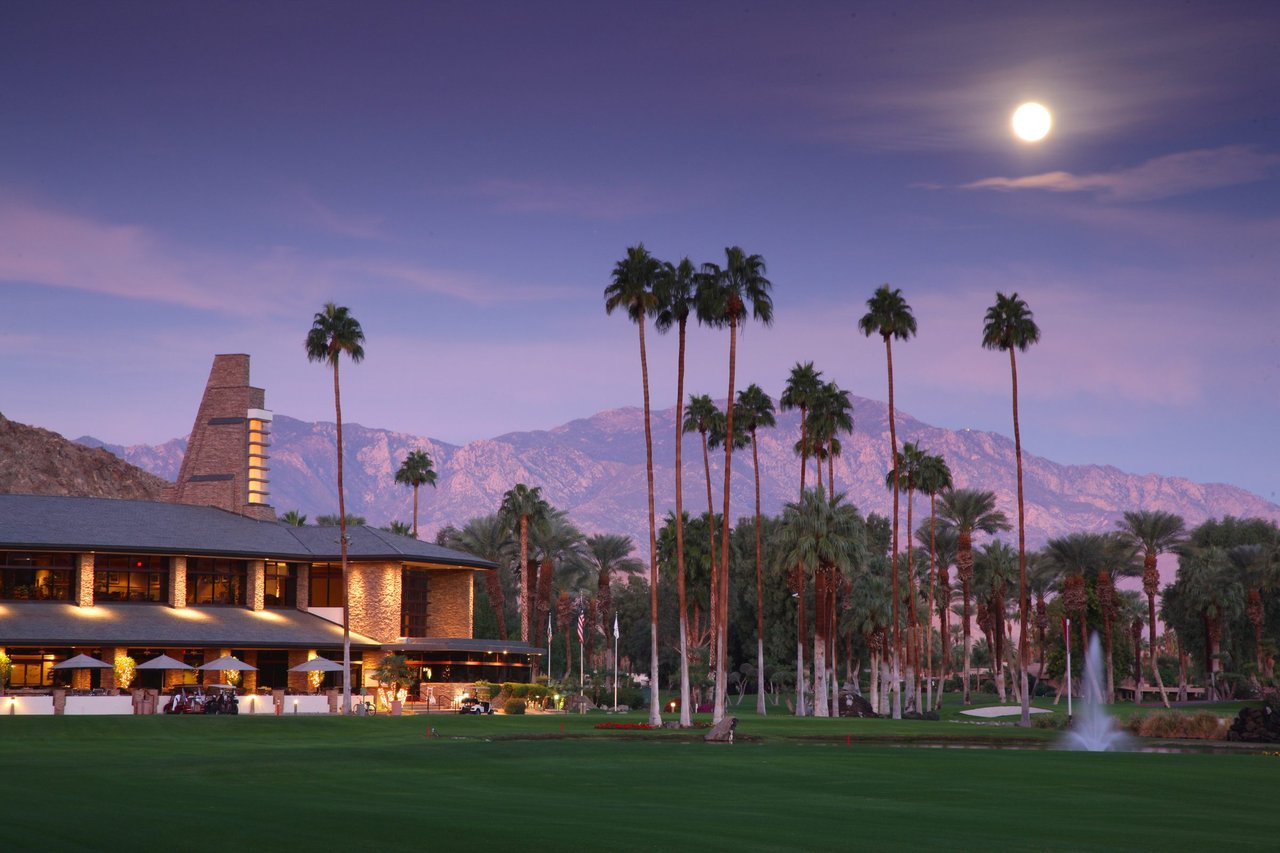 Indian Wells Country Club clubhouse at dusk, stone chimney and palms beside a fairway with the Santa Rosa Mountains behind