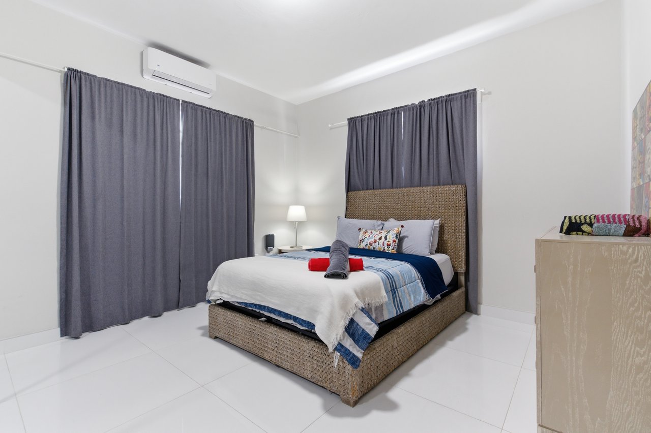 Furnished bedroom with a woven bed, blue-and-white bedding, gray curtains, and a wall-mounted air conditioner.
