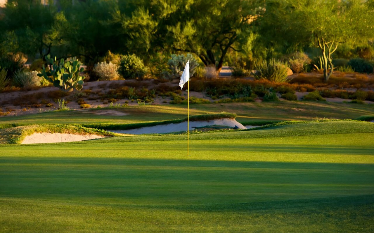 Golf green with a white flagstick, sand bunkers, and desert vegetation in golden light.