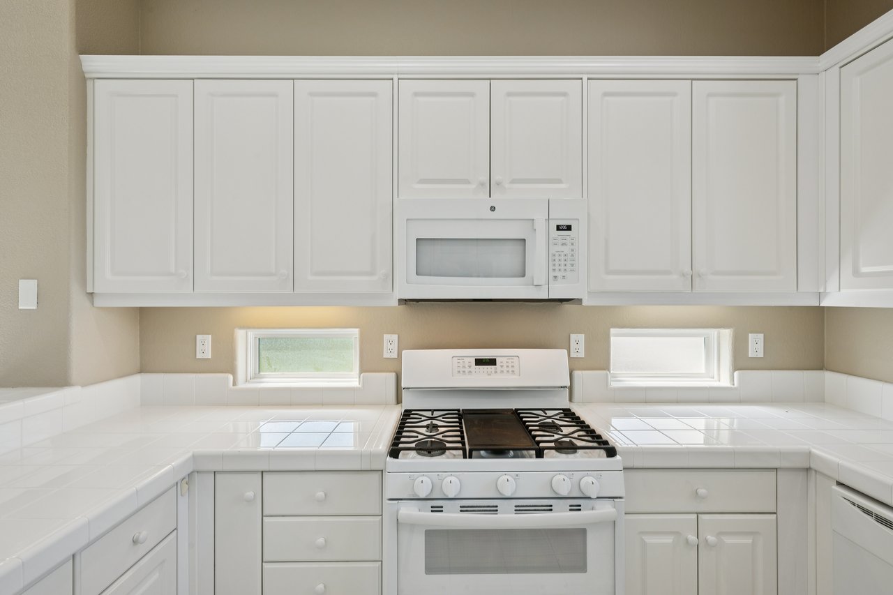 White kitchen with tiled counters, extensive cabinetry, and a gas range beneath a built-in microwave.
