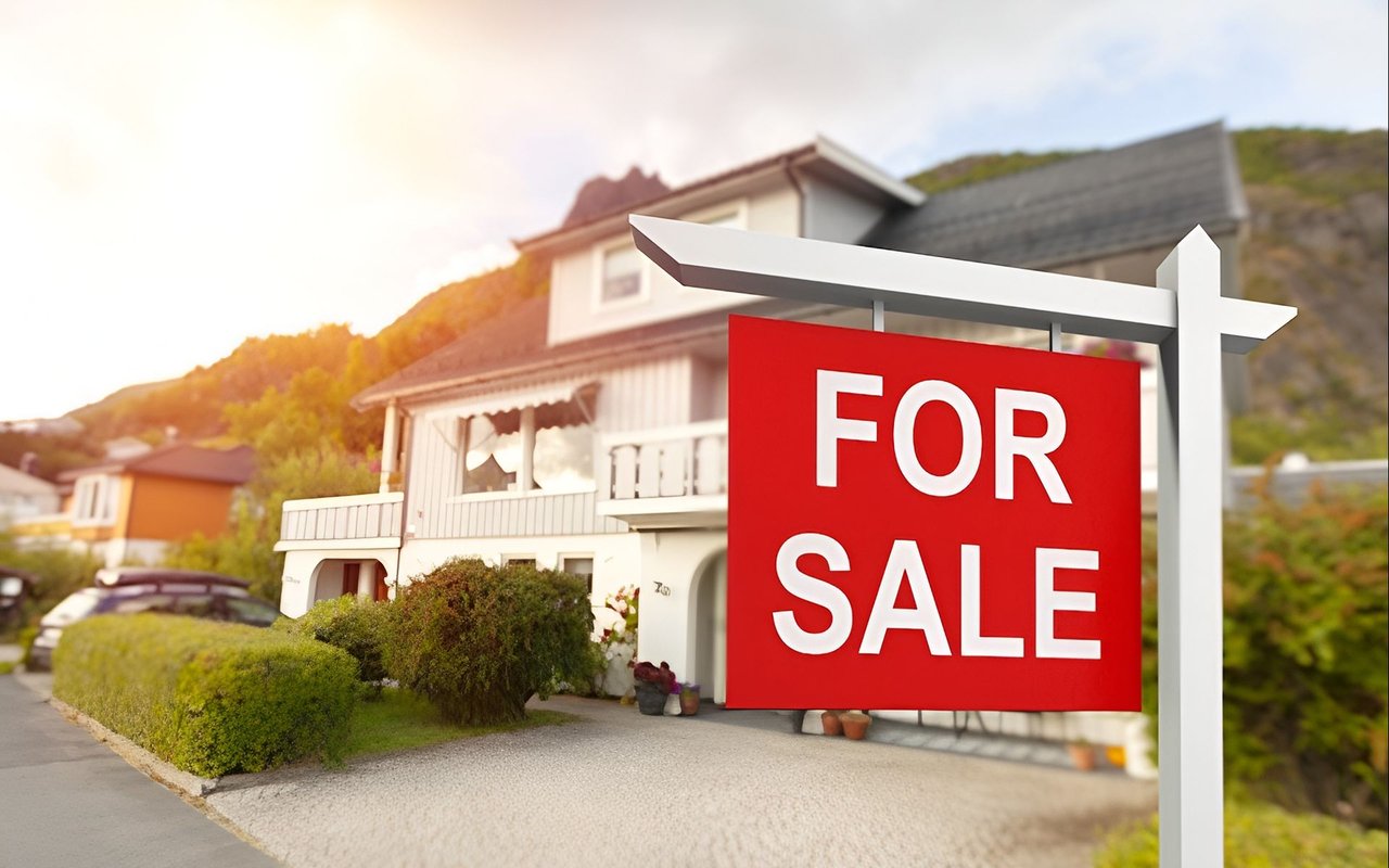 White two-story house with a balcony behind a prominent red FOR SALE sign.