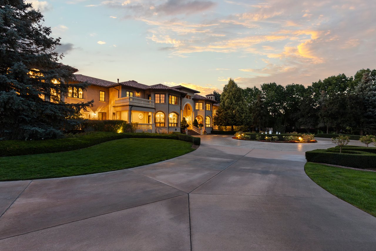 Mediterranean-style estate with warmly lit windows, landscaped grounds, and a curved driveway at sunset.