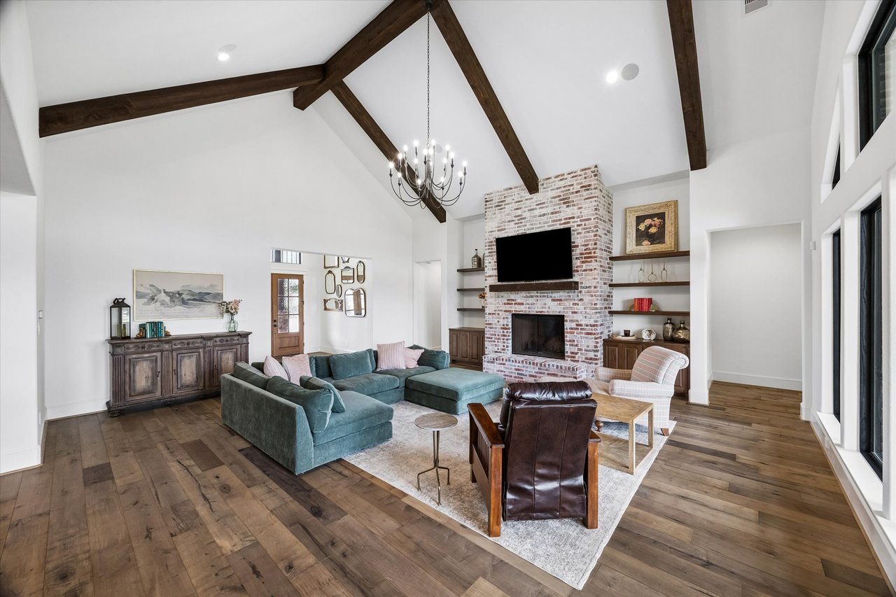 Living room with vaulted wood-beamed ceiling, brick fireplace, built-in shelves, and large windows.
