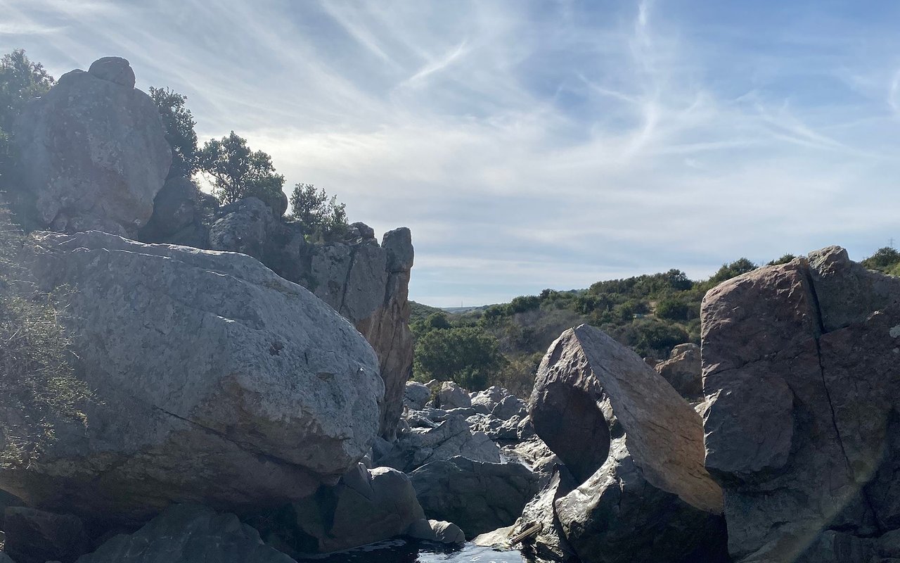 Large boulders frame a scrub-covered hillside beneath a cloudy blue sky.