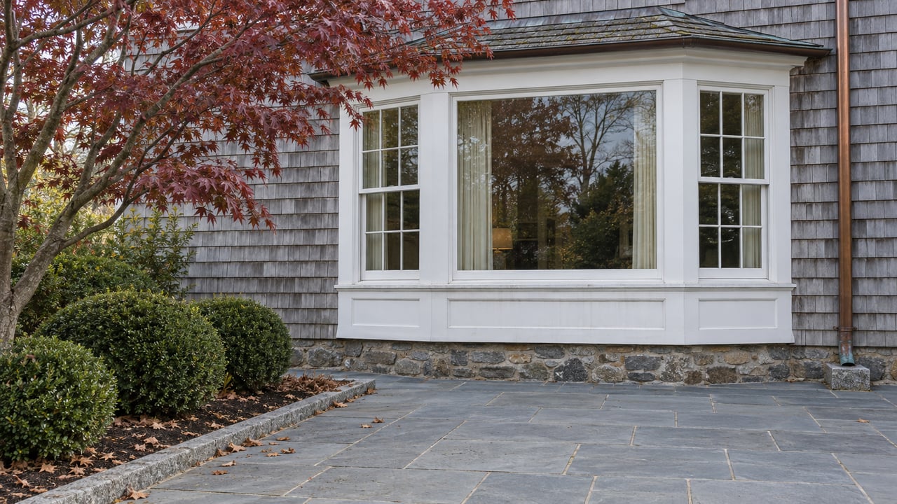A white-trimmed bay window in weathered cedar shingles above a bluestone terrace, framed by burgundy maple leaves and clipped inkberry.