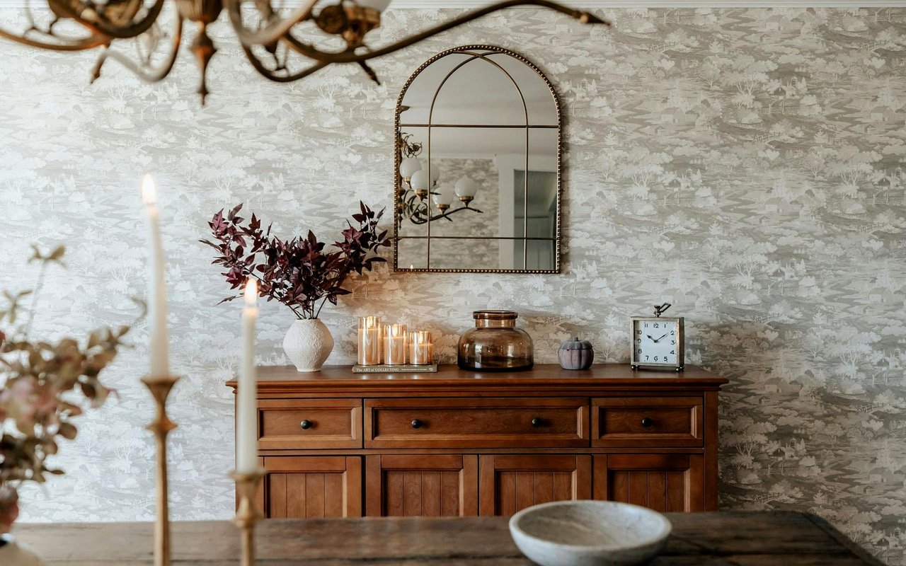 Furnished interior with a dark wood sideboard, arched mirror, patterned wallpaper, and candlelit decor.