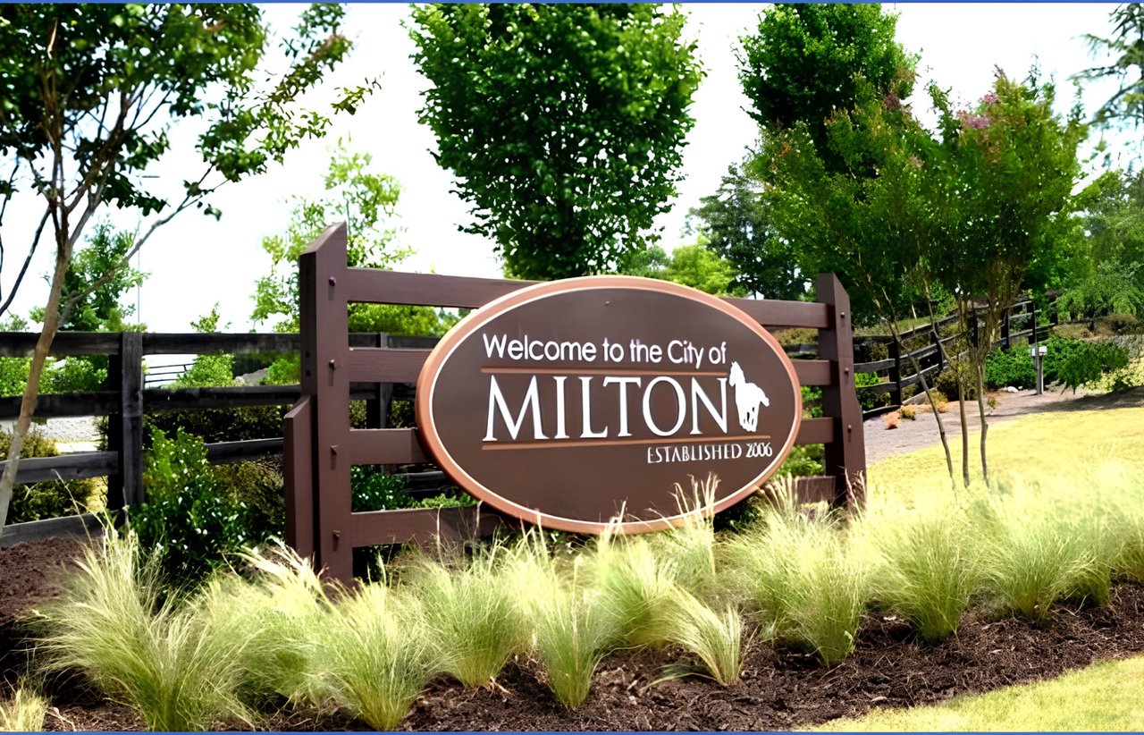 Milton welcome sign among trees and ornamental grasses.
