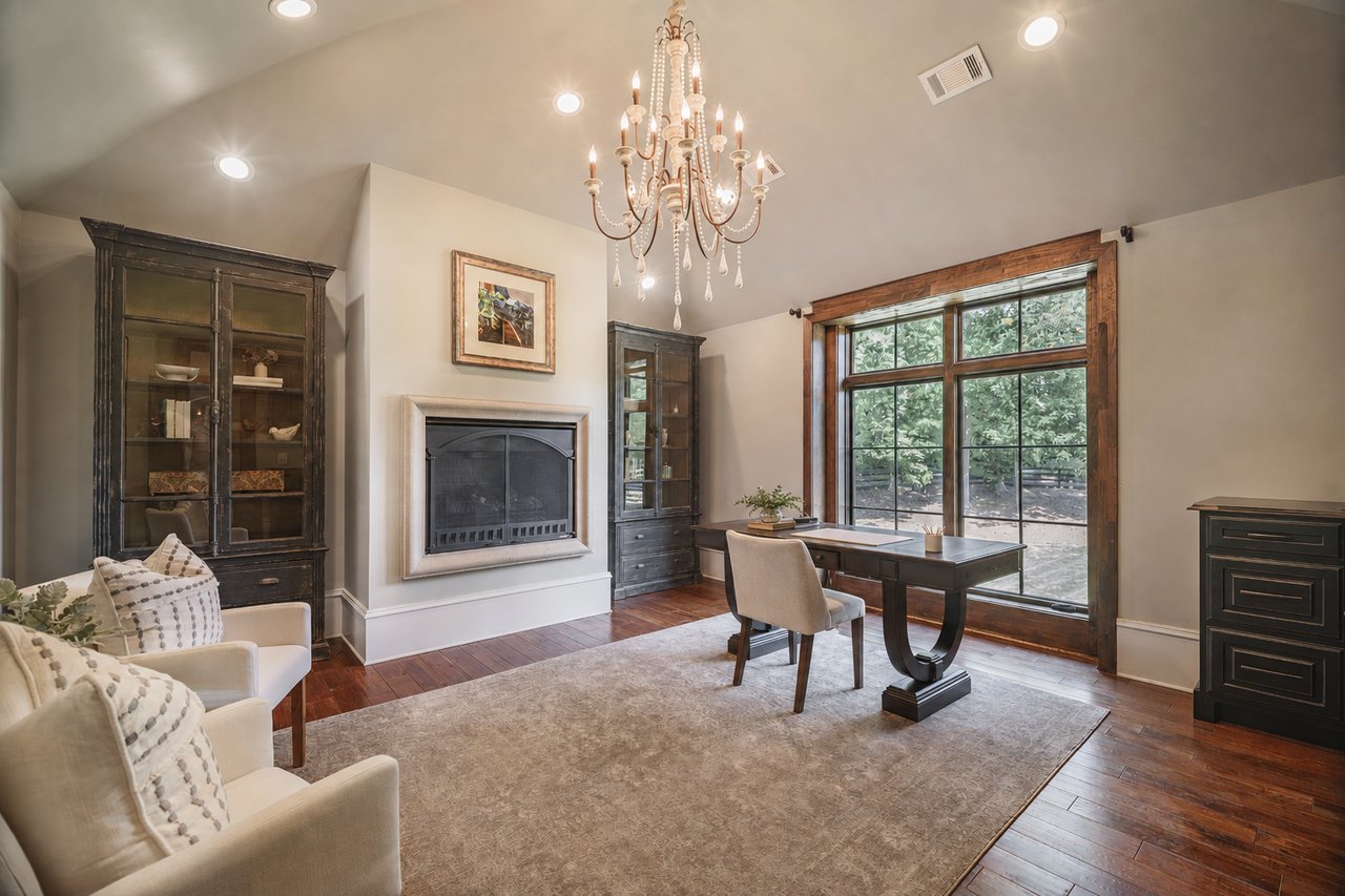 Furnished living room with a fireplace, desk, dark built-in cabinets, and large wood-trimmed window.