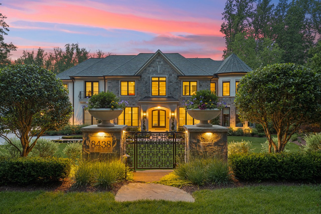 Stone-fronted residence with illuminated windows, landscaped entrance, and ornate gate at dusk.