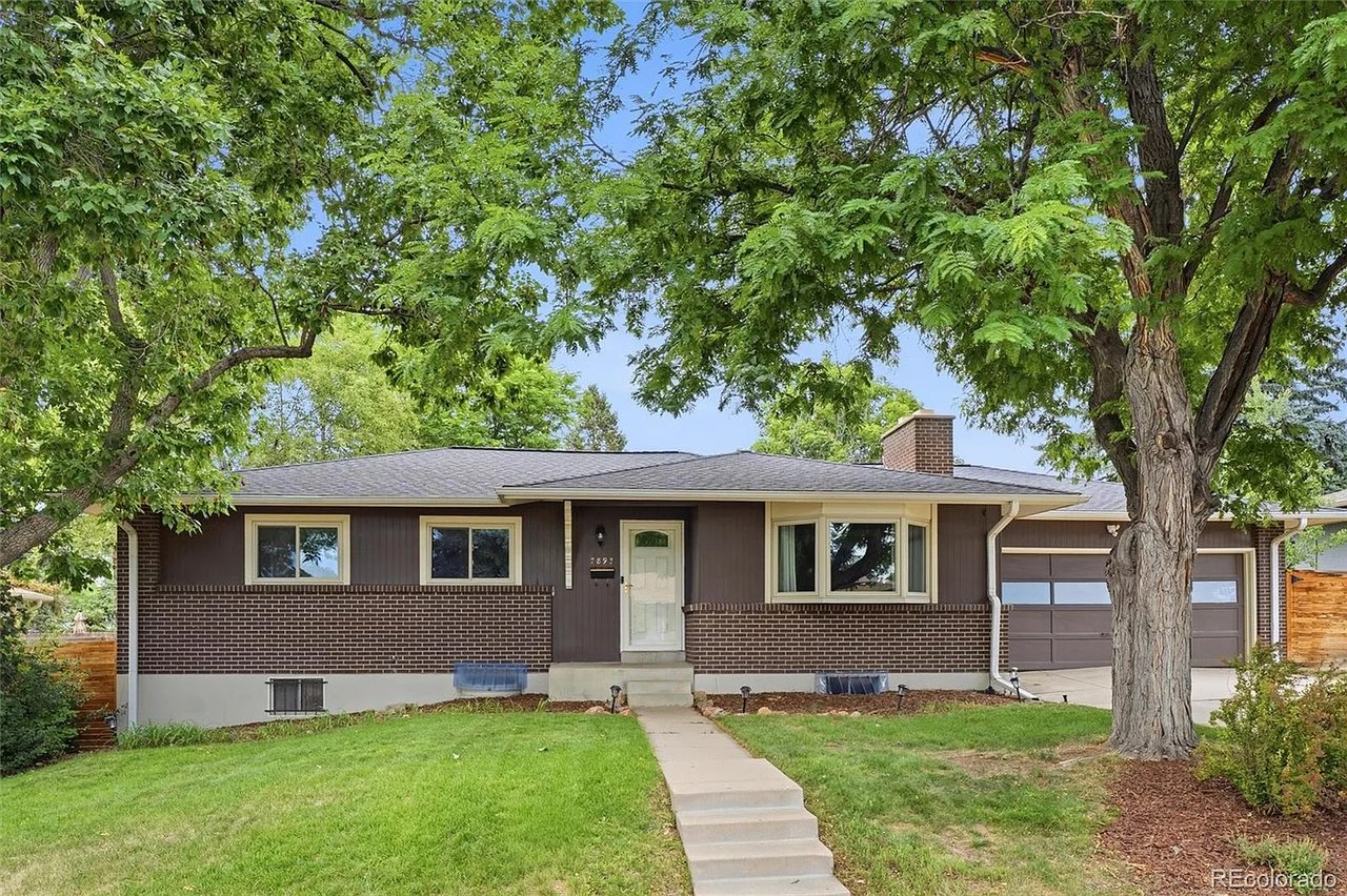 Single-story brick-and-siding home framed by mature trees, with a front lawn, walkway, and attached garage.