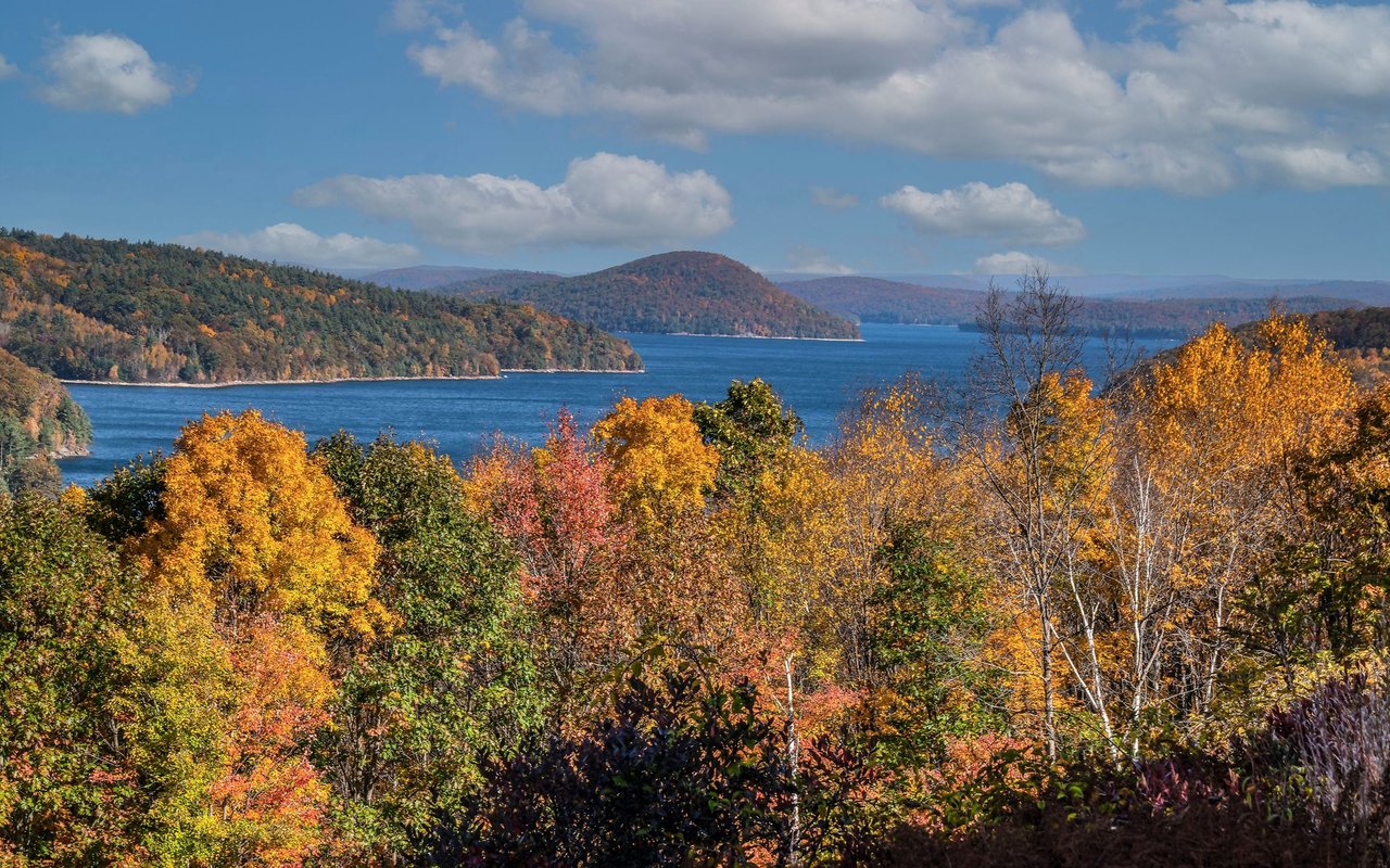 Autumn foliage frames a broad blue lake and forested hills beneath a partly cloudy sky.