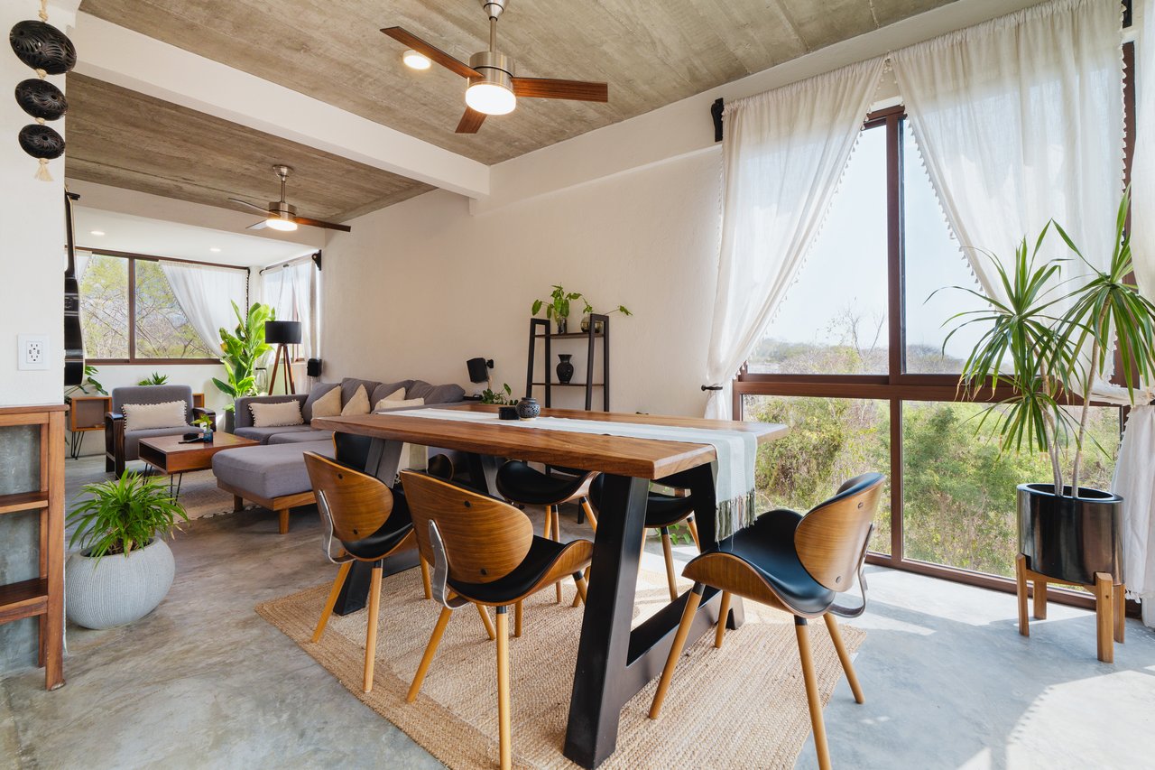 Open-plan living and dining room with a long wooden table, exposed concrete ceiling, and large windows overlooking greenery.