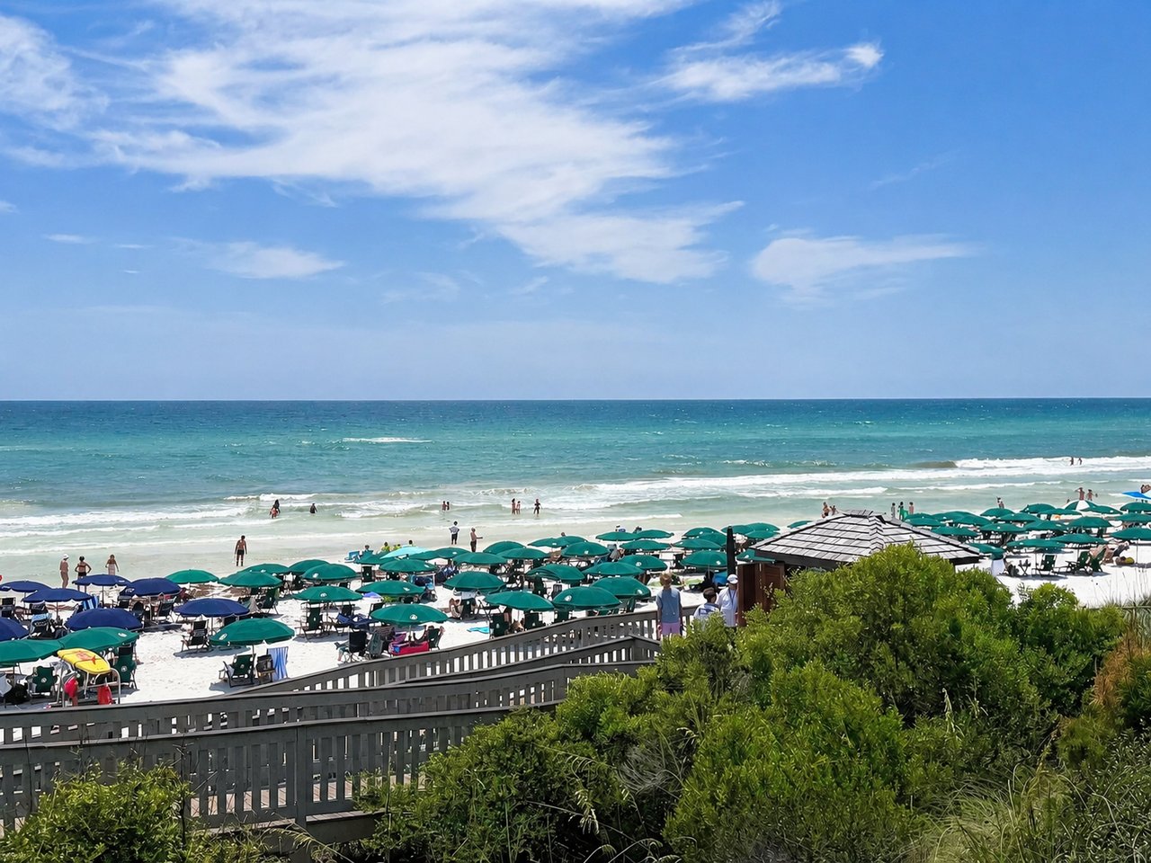 A scenic view from a wooden boardwalk looking over green coastal bushes toward a white sand beach filled with green patio umbrellas and the ocean under a partly cloudy blue sky.