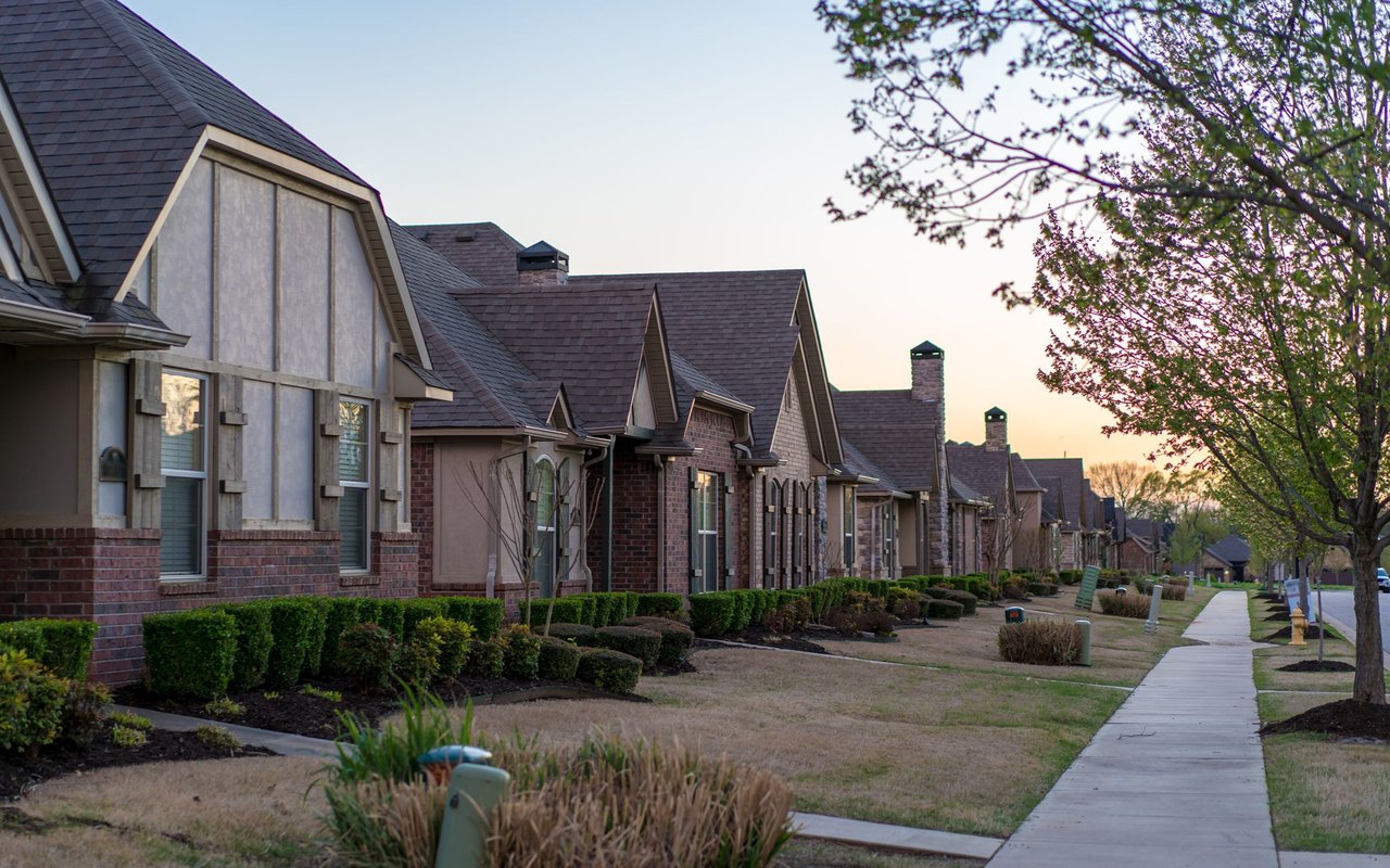 Suburban homes line a sidewalk, with brick and timber-style facades and neatly trimmed front gardens.