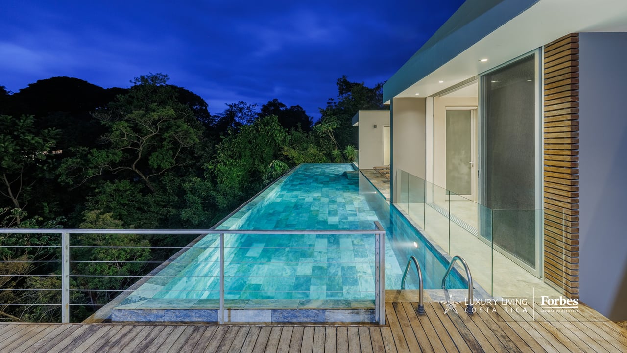 Modern home beside a long illuminated pool, with tropical forest beyond the glass railing.
