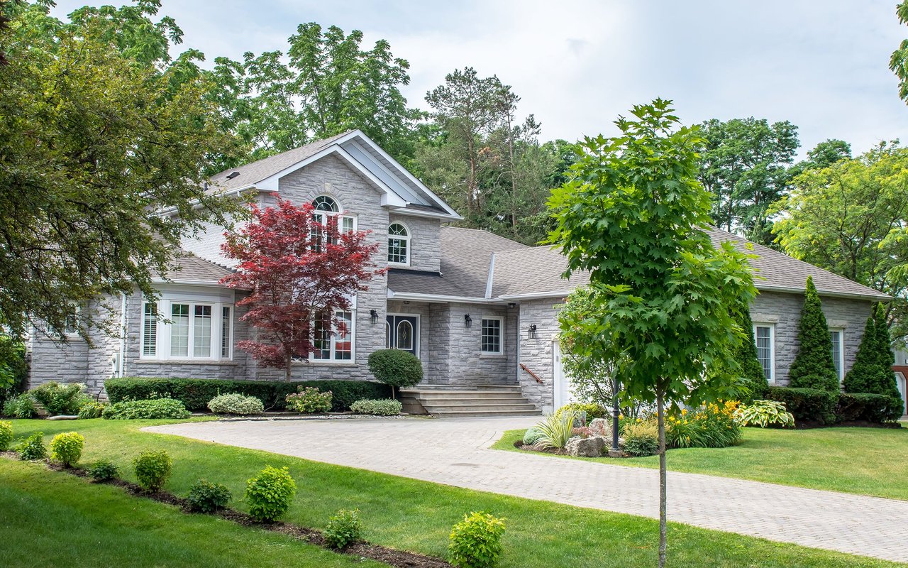 Gray stone house with arched windows, a paved driveway, and mature landscaped grounds.