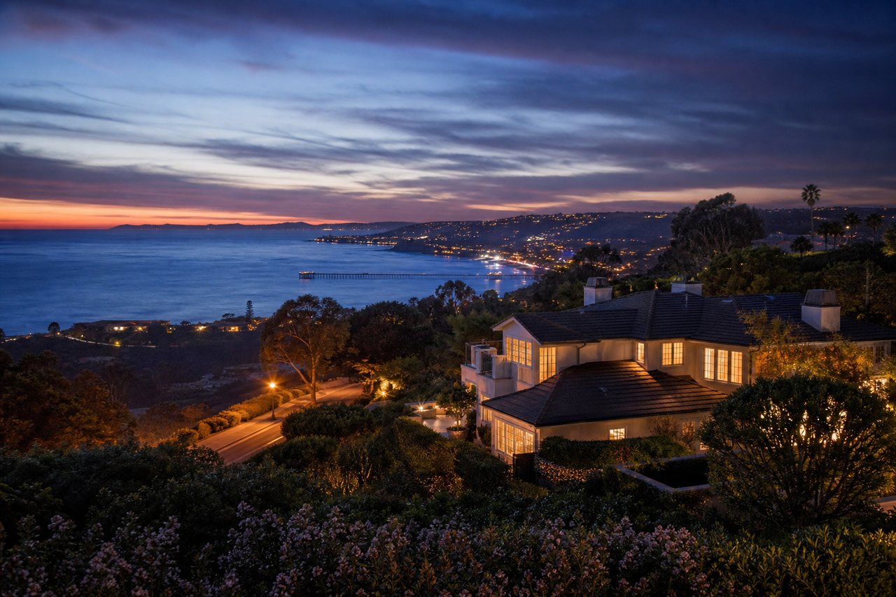 Hillside coastal home overlooking the ocean and illuminated shoreline at dusk.