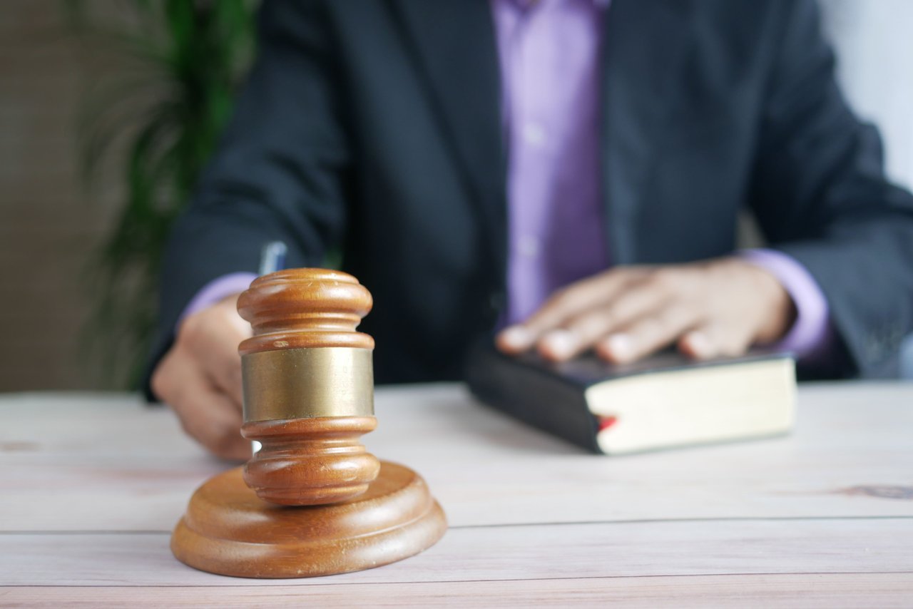 A wooden judge’s gavel rests on a desk, with a suited person and book blurred behind it.