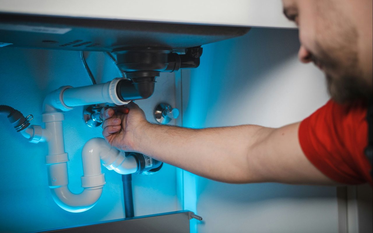 Plumber adjusting a fitting beneath a sink among white drain pipes.
