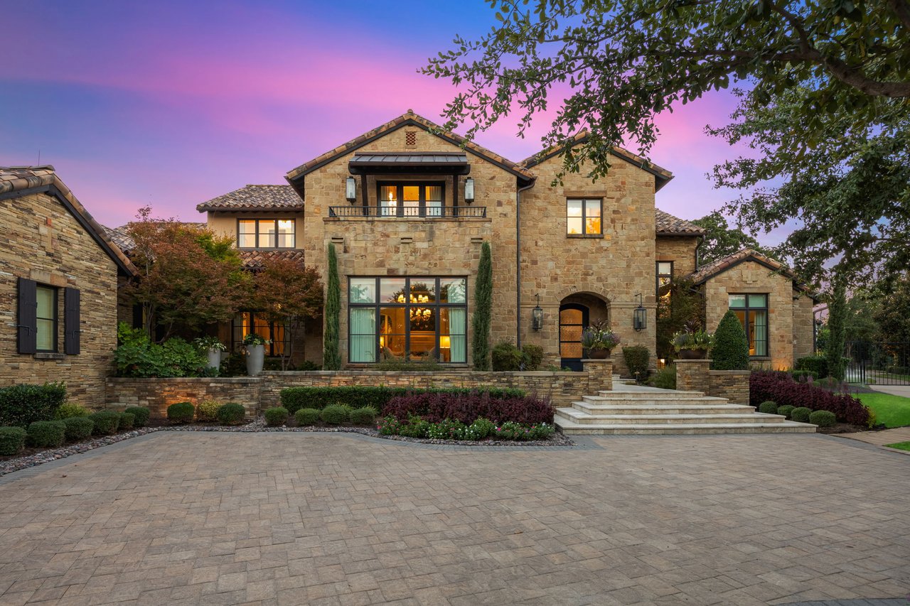 Two-story stone home with a broad paved driveway and landscaped front garden at dusk.