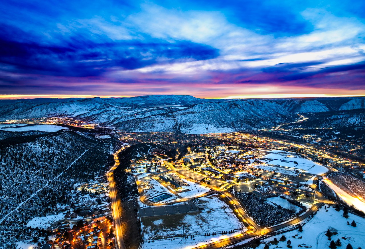 Snow-covered mountain town at dusk, with glowing streets and dramatic blue clouds.