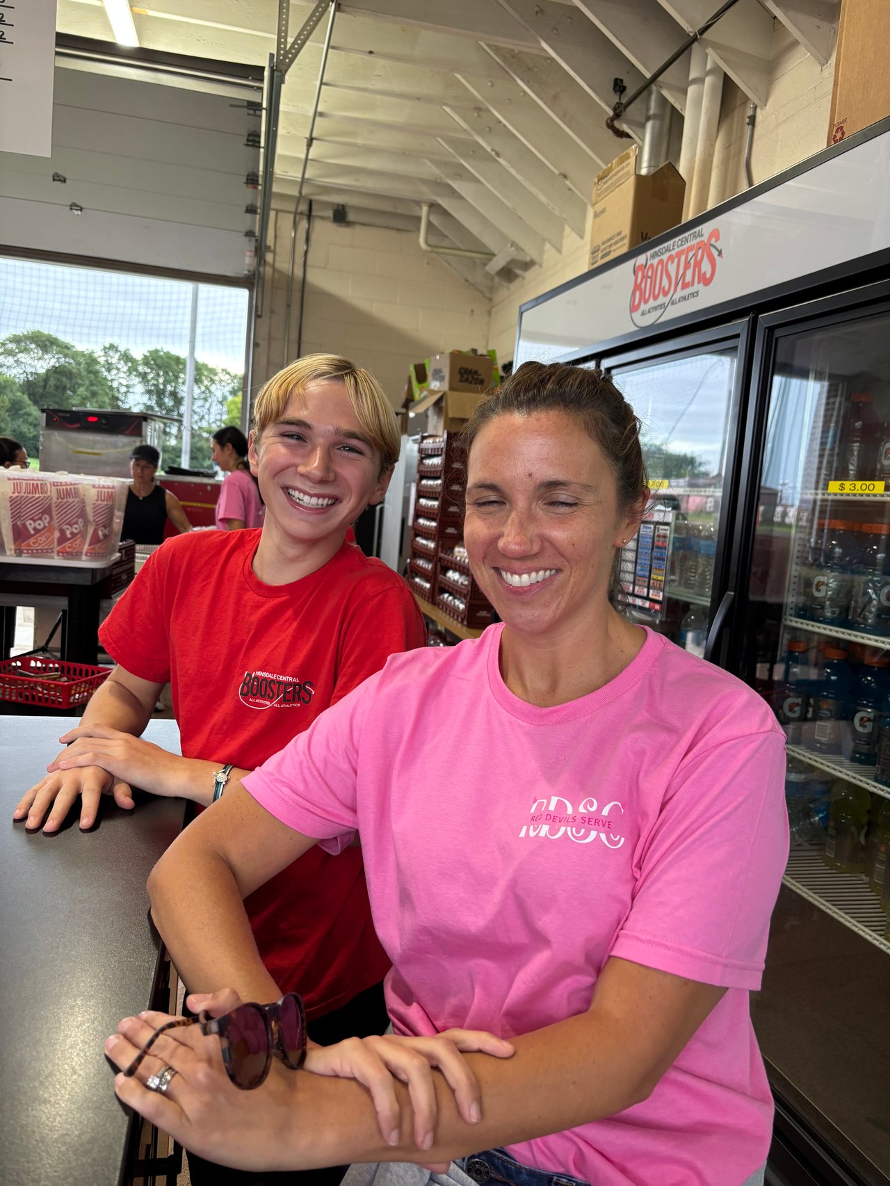 A teenage boy and woman smile at a concession counter in Hinsdale Central Boosters shirts.