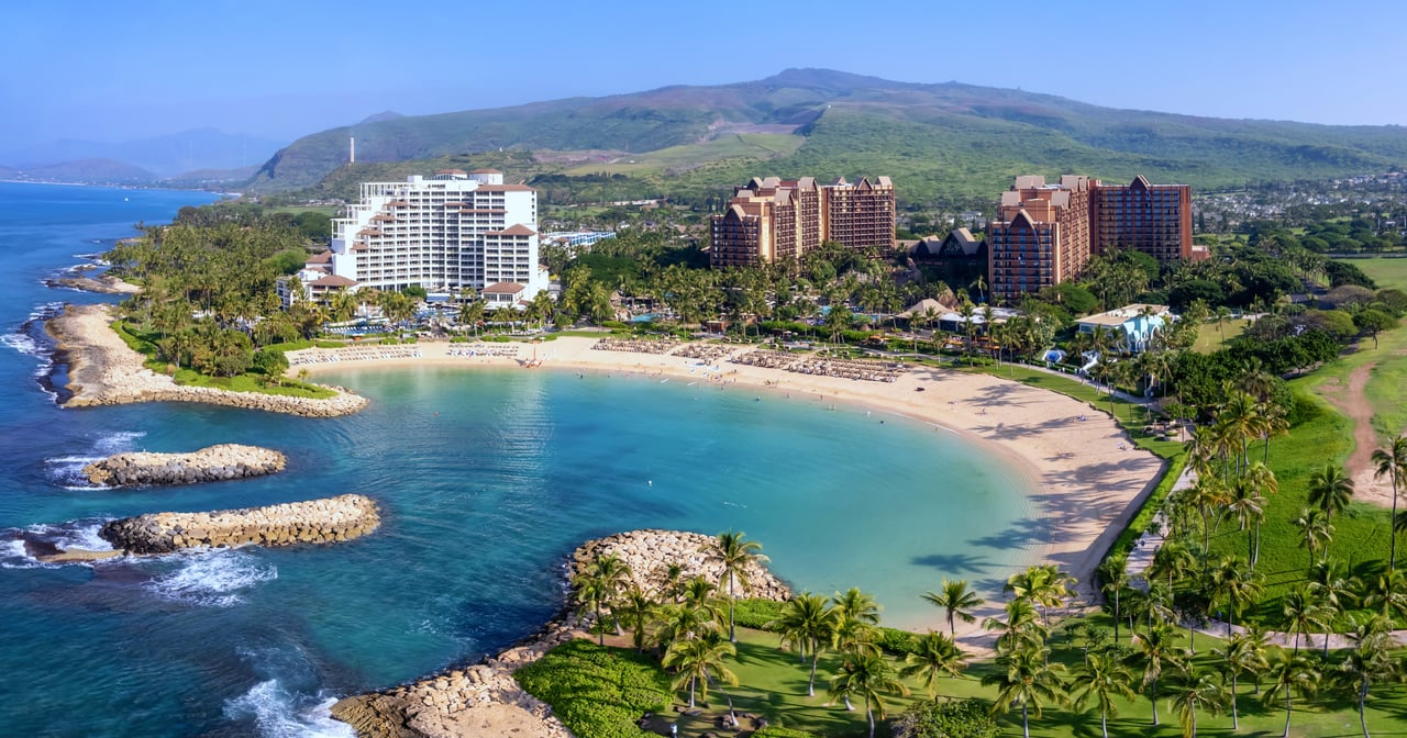 Curved sandy beach and turquoise lagoon bordered by palm-lined resort grounds and green mountains.