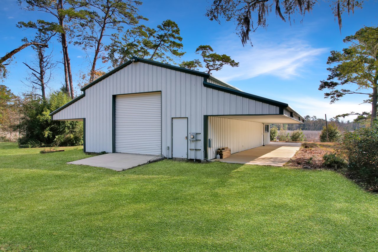 A metal garage with a white roll-up door is set in a lush green yard, surrounded by tall trees under a clear blue sky, conveying a peaceful, rural vibe.