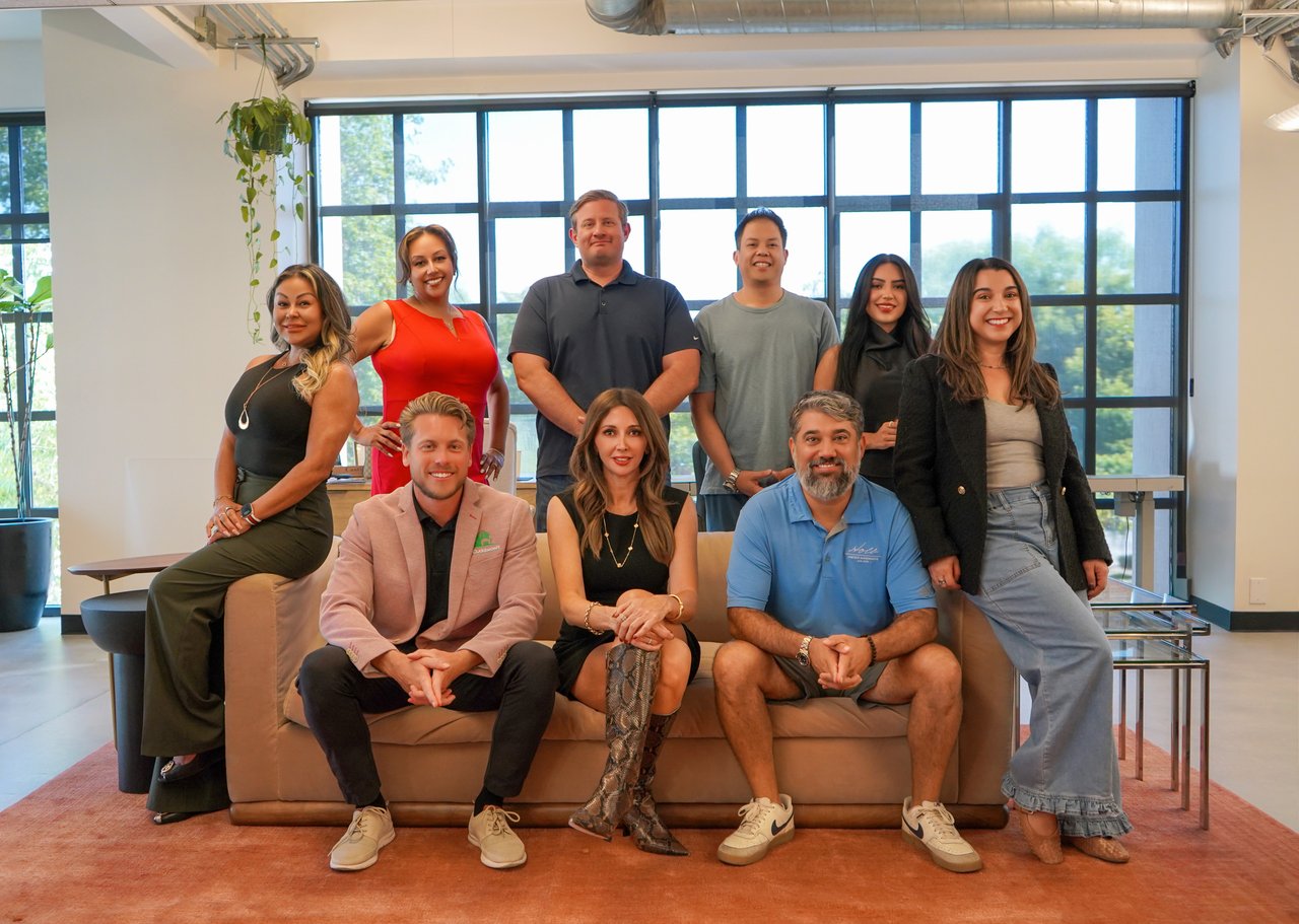 Nine real estate team members pose together in a bright office, with three seated on a sofa.